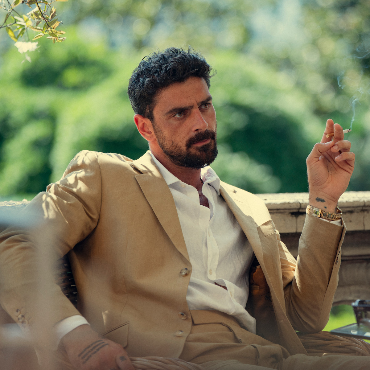 Man in a tan suit sitting outdoors on a patio, holding a cigarette, looking serious with greenery and sunlight in the background.