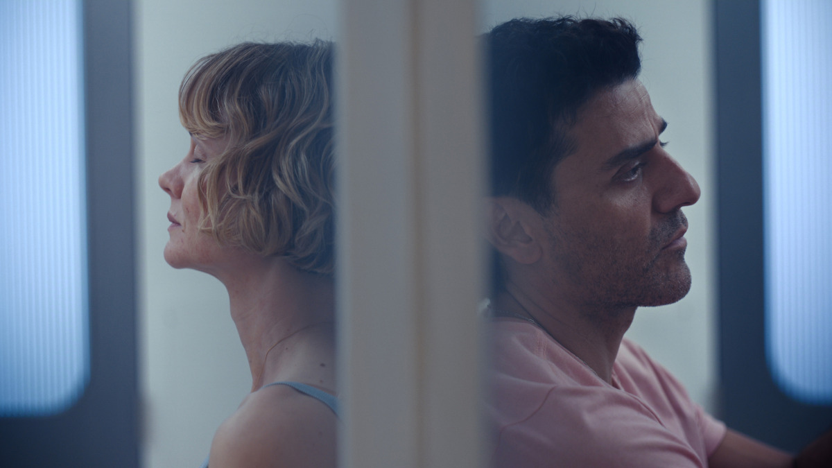 Two people sit back to back separated by a translucent barrier, both appear contemplative and distant, in a softly-lit modern environment with blue and white tones suggesting an emotional or dramatic scene.