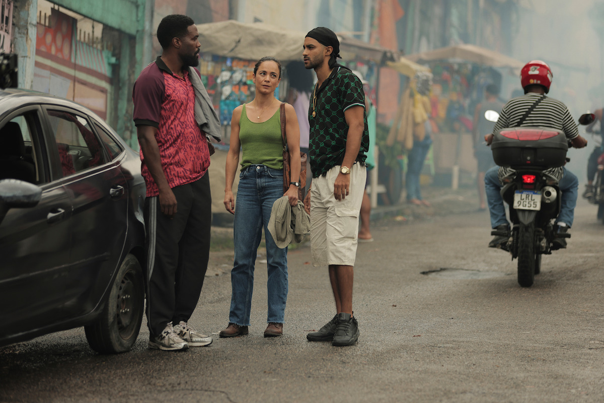 Three people stand talking on a busy, colorful street near a black car as a motorcyclist rides by. Market stalls and buildings line the background, suggesting an urban outdoor setting.