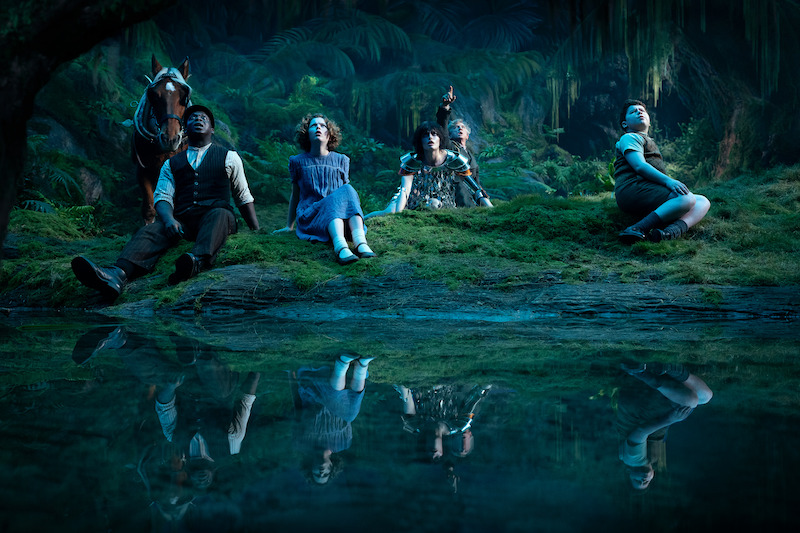 Five people and a horse sit on mossy ground beside a reflective pond in a mystical, lush forest at night, gazing upwards in wonder under blue atmospheric lighting.