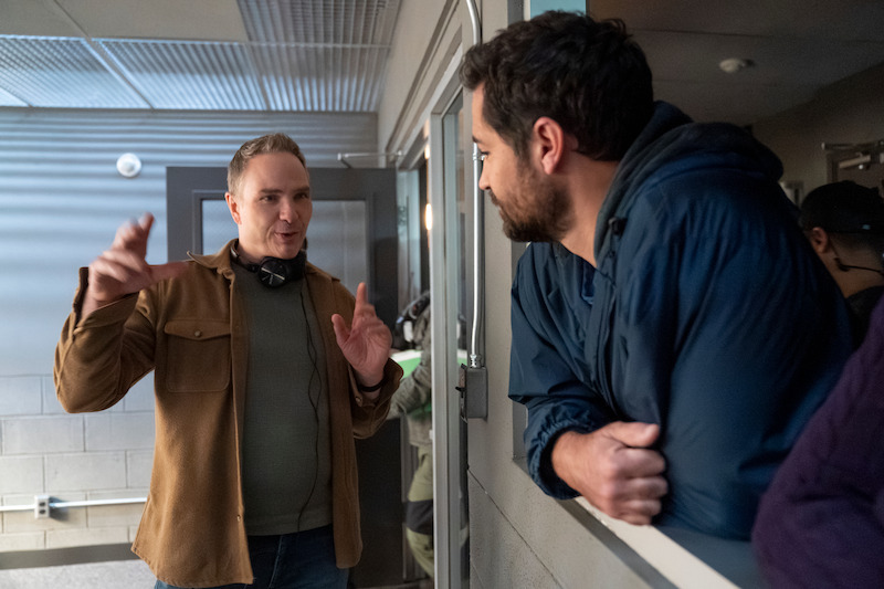 Two men have an animated conversation in a modern, industrial-looking room with gray walls and fluorescent lighting; one gestures while the other listens, creating a collaborative and focused mood.