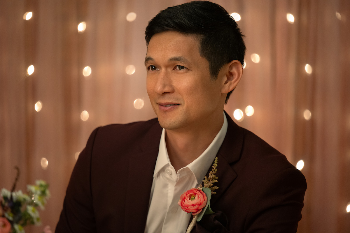 Man in a maroon suit with a boutonnière, seated indoors with warm string lights and soft brown drapery in the background, smiling at an event or celebration.