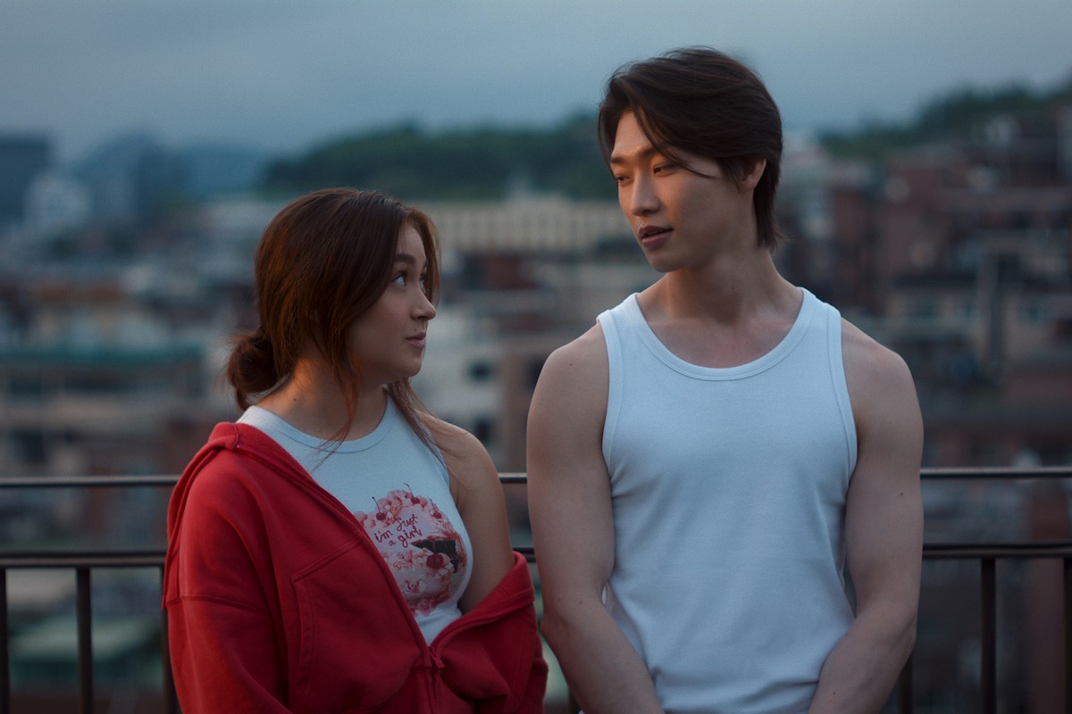 Two young adults talking on a rooftop at dusk, with city buildings and hills in the background; one wears a white tank top and the other a red jacket, capturing a relaxed, intimate urban setting.