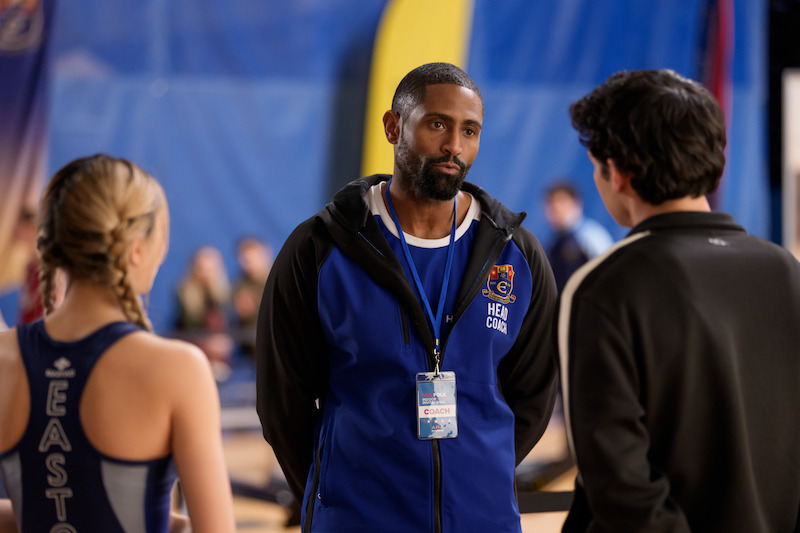 A coach in a blue and black jacket stands in a gymnasium talking to two athletes, one male and one female, with sports banners and people blurred in the background.