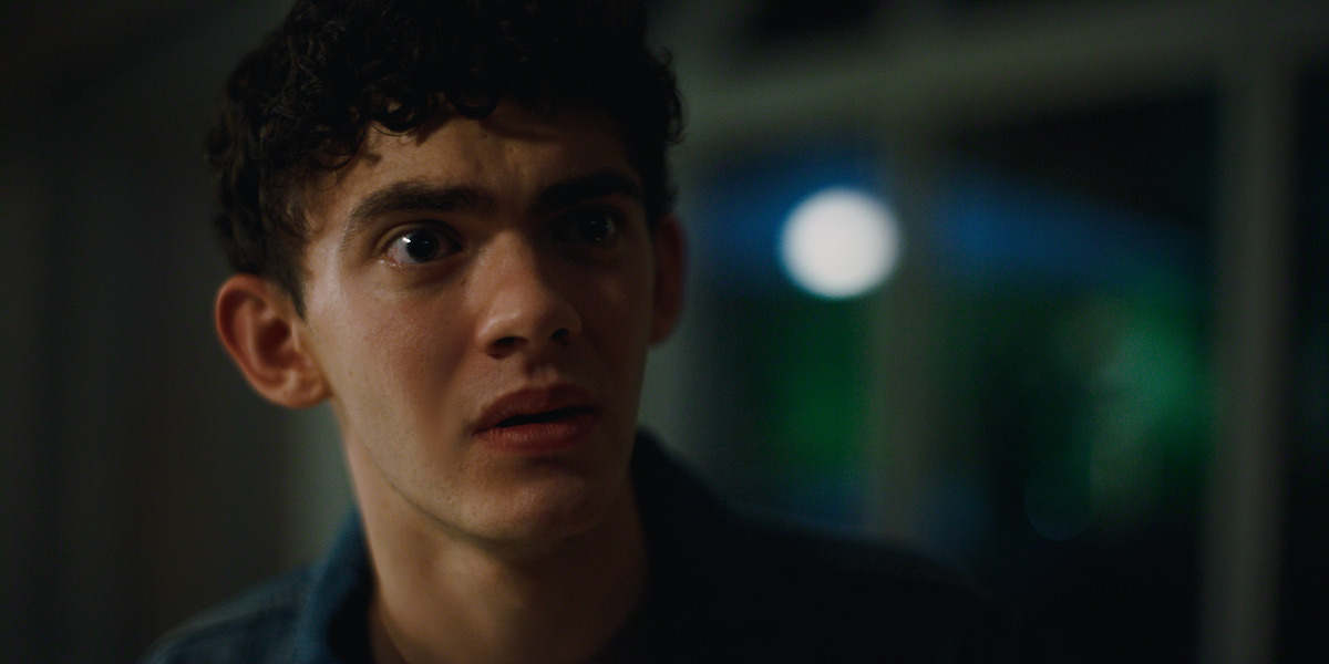 Young man with curly hair looking surprised or anxious indoors at night, with blurred windows and bright light visible in the background.