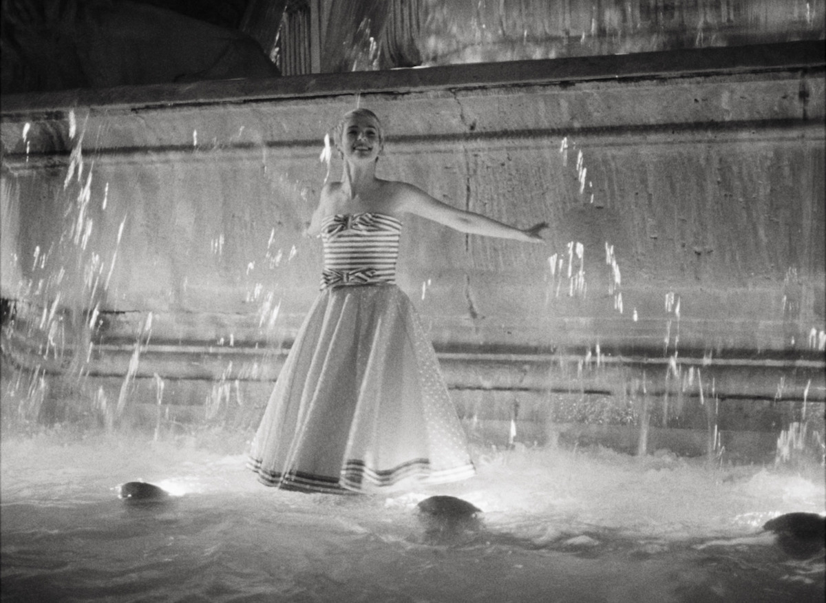 Zoey Deutch in a strapless dress stands in a fountain in a film still from Nouvelle Vague