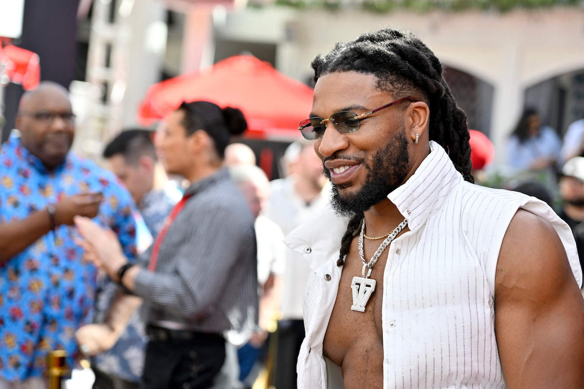 Smiling man with dreadlocks, glasses, and jewelry in a sleeveless vest at an outdoor event, surrounded by people and red umbrellas in the background.