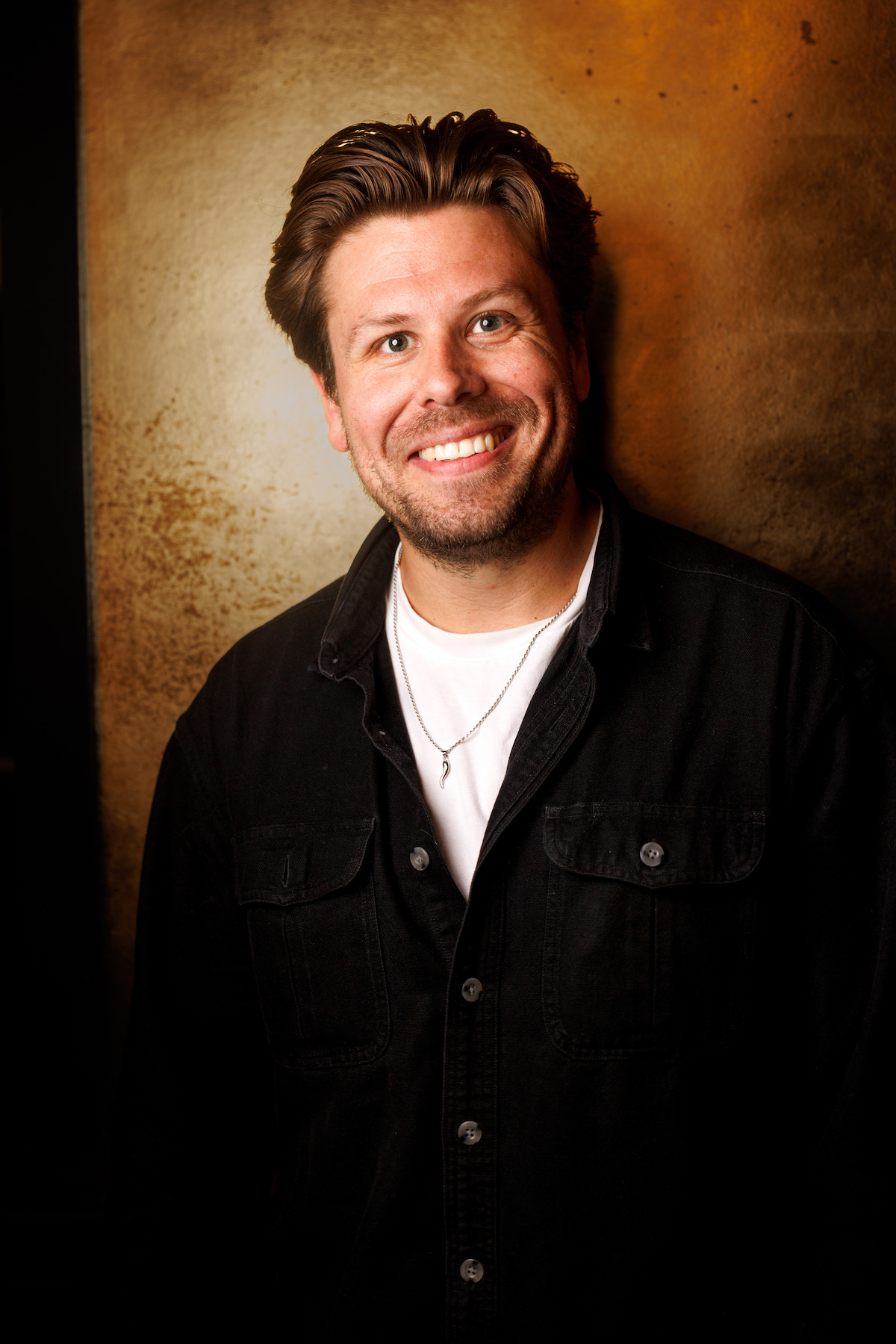 Smiling man with styled brown hair wearing a black shirt and white t-shirt, standing indoors against a warm, textured brown and gold background, in relaxed and casual pose.