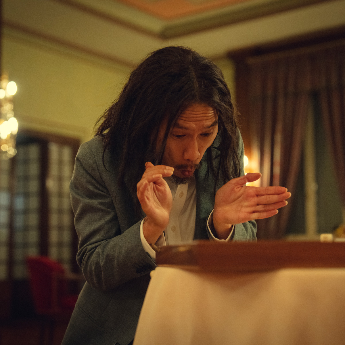 Man with long hair and formal jacket leans over a table, making an expressive gesture with hands in an elegant, warmly lit room with curtains and chandeliers in the background.