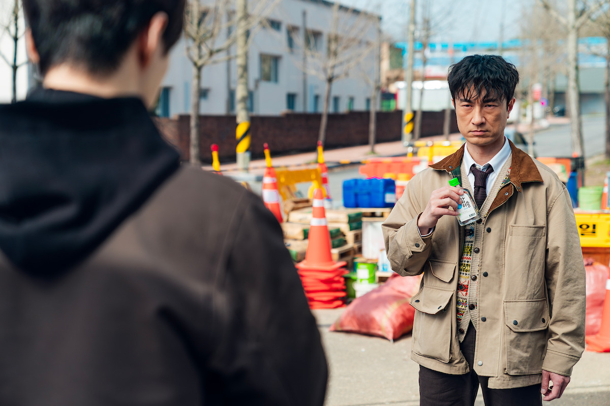 A serious man in a tan jacket holds a drink, standing outdoors at a construction site with traffic cones and bags, facing another person on a chilly, overcast day, creating a tense urban atmosphere.