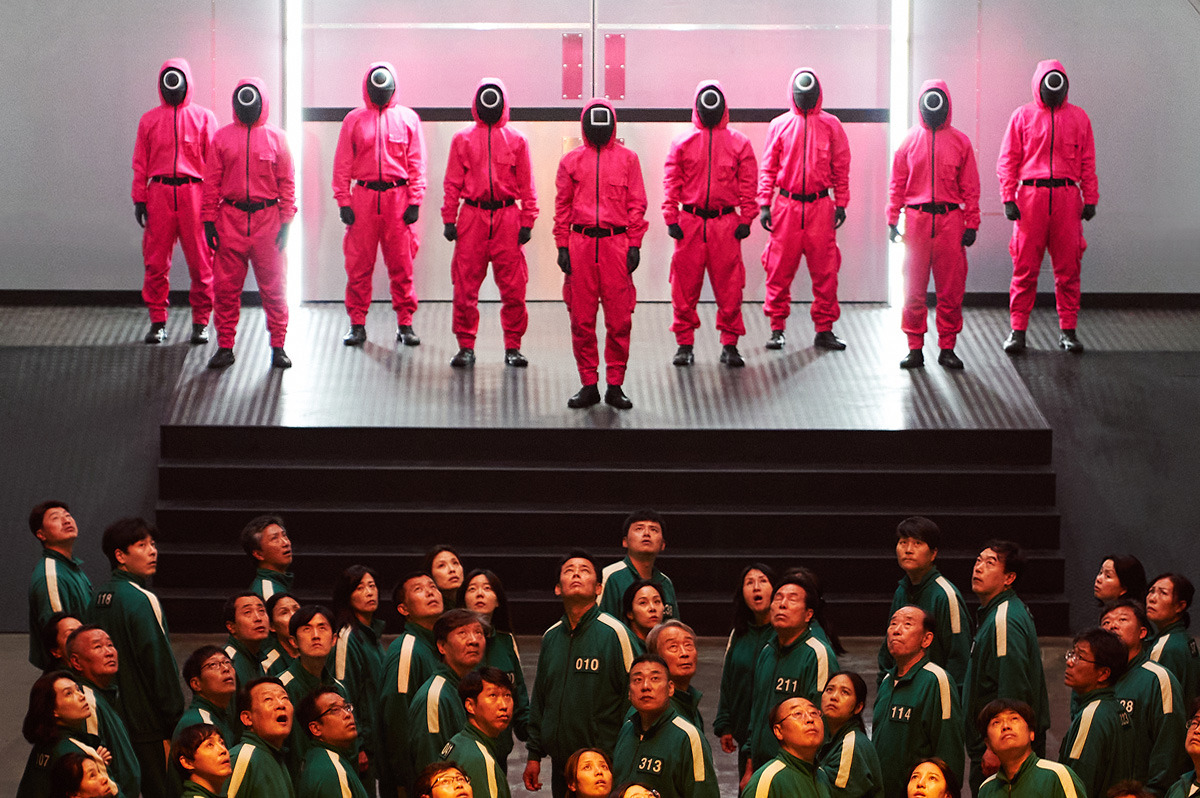 Instream still from season one of 'Squid Game', showing one group wearing the iconic pink jumpsuits and another group sporting the green and white tracksuits.