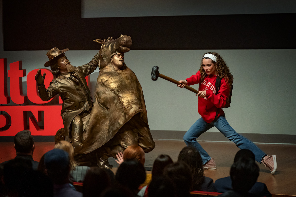 A young woman in a red sweater and jeans wields a sledgehammer on stage, aiming at a bronze statue of a cowboy and a bull, in front of an audience during a presentation or performance.