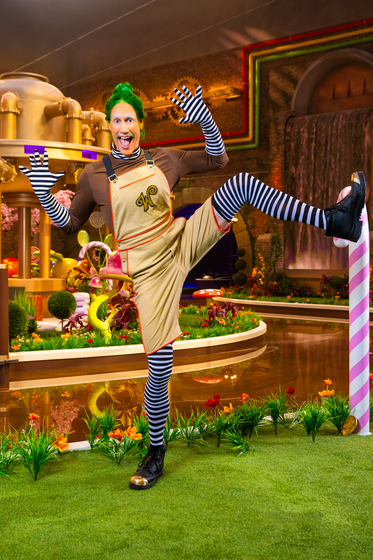 A person dressed as an Oompa Loompa with green hair, striped stockings, and colorful costume poses playfully in a whimsical, candy-themed environment with plants, water, and vibrant decorations.