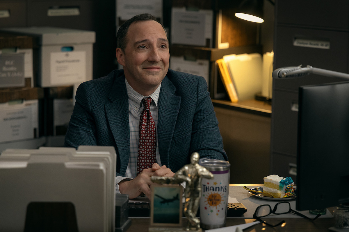 A man in a suit sits smiling at a desk in an office, surrounded by files, folders, a cake slice, a trophy, and a coffee mug, with shelves and organized paperwork in the background under warm lighting.