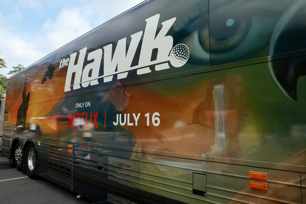 A bus with a colorful mural for a Netflix show called "The Hawk," featuring large hawk imagery, silhouettes, and a golf ball. The environment is an outdoor parking area with part of another vehicle visible in the background.
