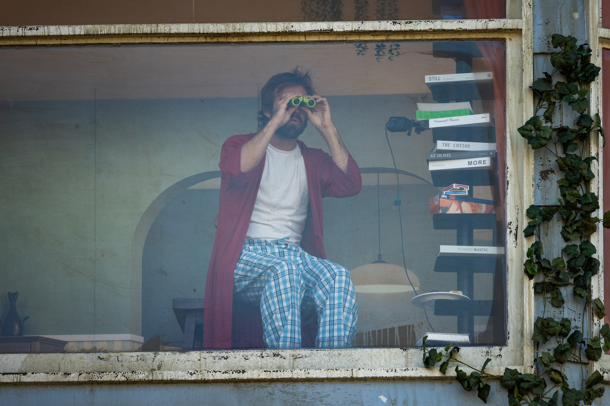 Man in pajamas and red robe sits inside, looking through binoculars out a large window. The indoor setting has stacked books, a lamp, and houseplants visible, with vines growing on the building’s exterior.