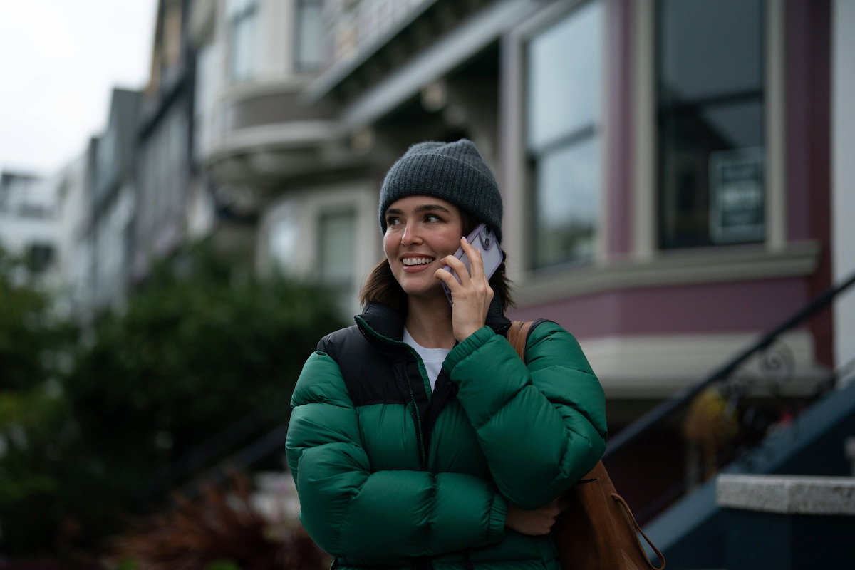 Woman in a green jacket and gray beanie smiling while talking on a phone outdoors in a residential urban neighborhood with houses and stairs in the background on a cloudy day.