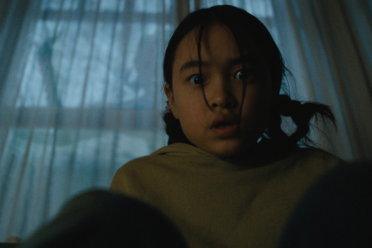 A young girl with pigtails sits indoors, looking surprised or scared, with wide eyes. She is in a dimly lit room with sheer curtains and a window showing a dark, moody outdoor scene in the background.