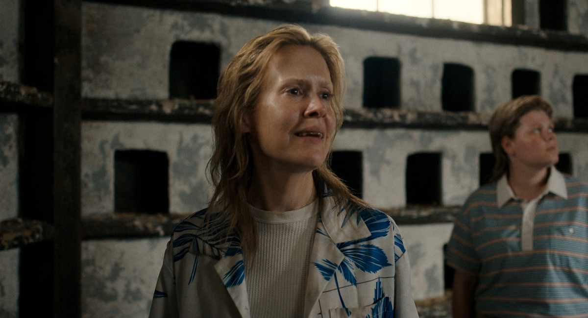 Two people stand indoors near a wall with square openings and weathered paint. The person in front wears a patterned shirt and looks ahead, while the second person glances off to the side in a striped polo.