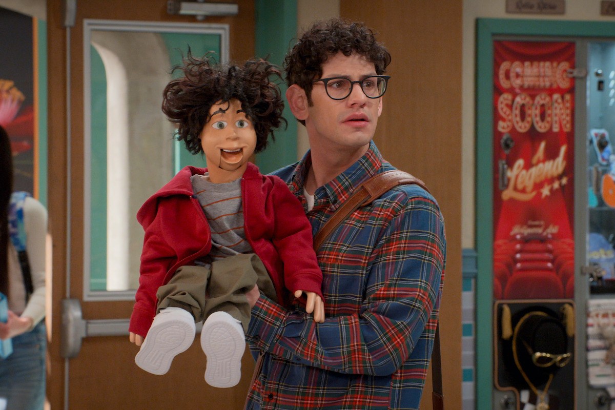 A young man with curly hair and glasses holds a ventriloquist dummy in a school hallway, standing near a vending machine and a door with posters and lockers visible in the background.