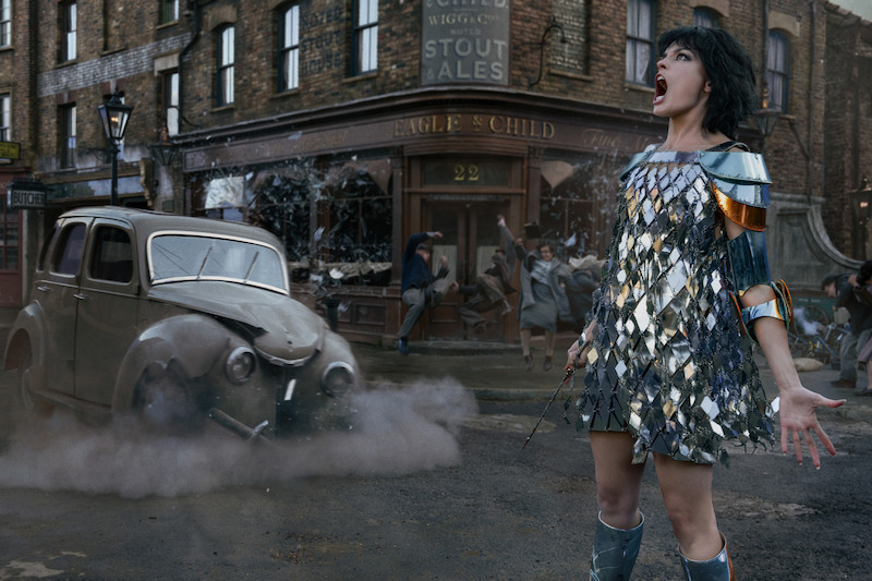 A woman in a shiny dress stands on a city street near an old car and a pub as chaos unfolds, with people running and a shattered window in the background, dust and debris filling the scene.