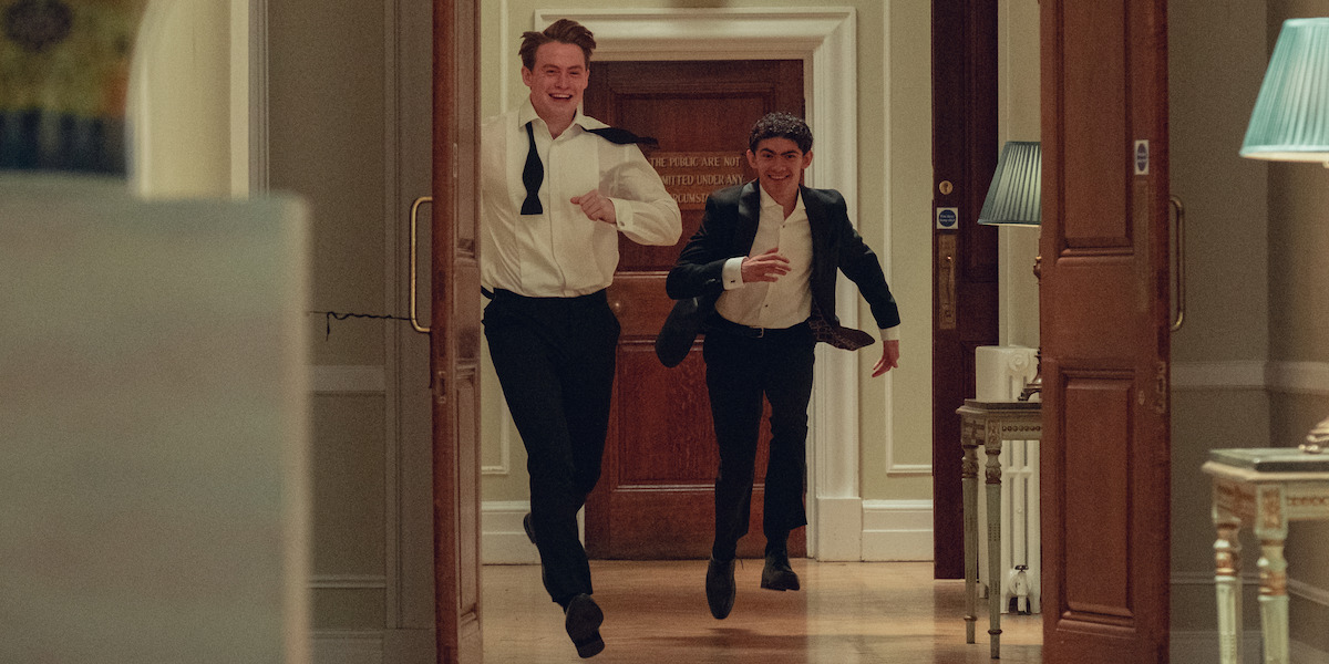 Two young men in suits and untied bow ties joyfully run down a well-lit hallway in an elegant building, appearing happy and lively. The environment features wooden doors, classic decor, and soft lamp lighting.