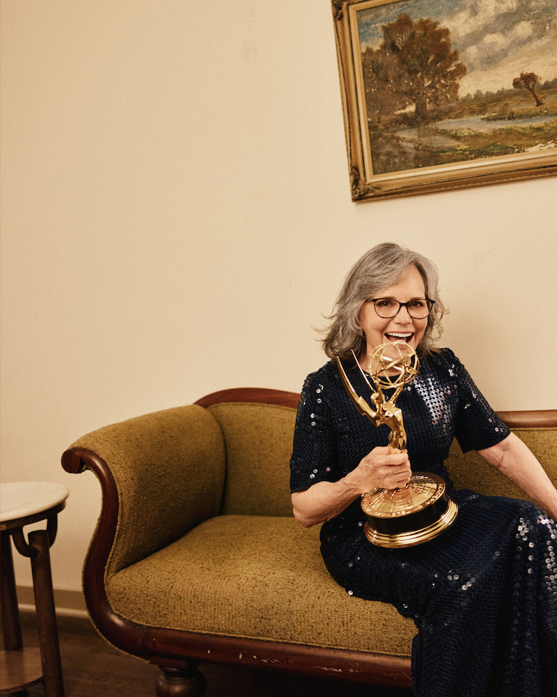 Smiling woman in a dark sequin dress sits on a vintage green couch, holding a large Emmy award. The setting is indoors with neutral walls, wooden floors, a small side table, and a framed landscape painting on the wall.