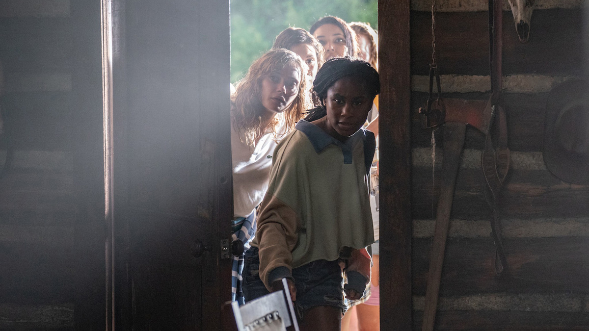A group of young women cautiously peek through a cabin door into a dimly lit rustic room with wooden walls and hanging tools, looking alert and tense.