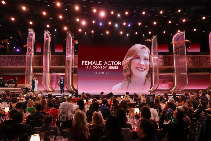 Award show scene with a large audience seated at tables, presenters on stage, and a big screen displaying "Female Actor in a Comedy Series - Television" with Catherine O'Hara's smiling portrait. Elegant venue with bright stage lights.