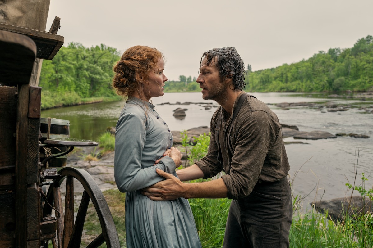 A man and woman in old-fashioned clothing stand by a river, holding each other by a wagon wheel in a lush, green outdoor setting, suggesting a historical or pioneer era moment filled with emotion.