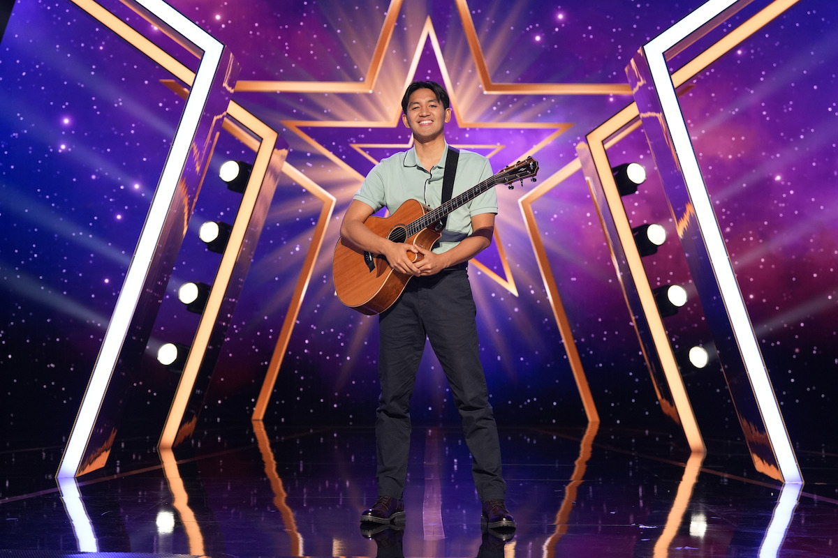 JR De Guzman stands center stage holding an acoustic guitar, surrounded by illuminated stage lights and a large glowing star backdrop on a starry, purple-themed talent show set.