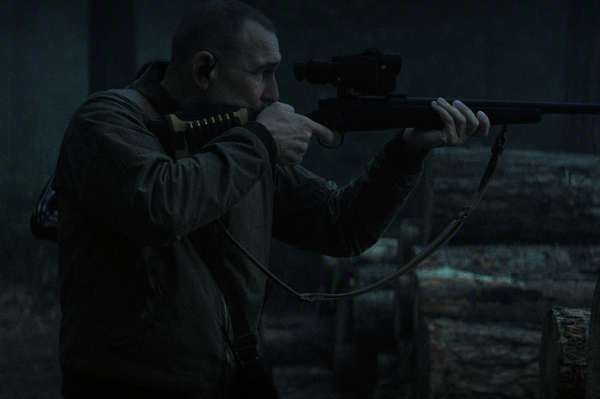 Man aiming a scoped rifle in a dark, outdoor forest setting with large logs in the background, wearing a brown jacket, focused on his target.
