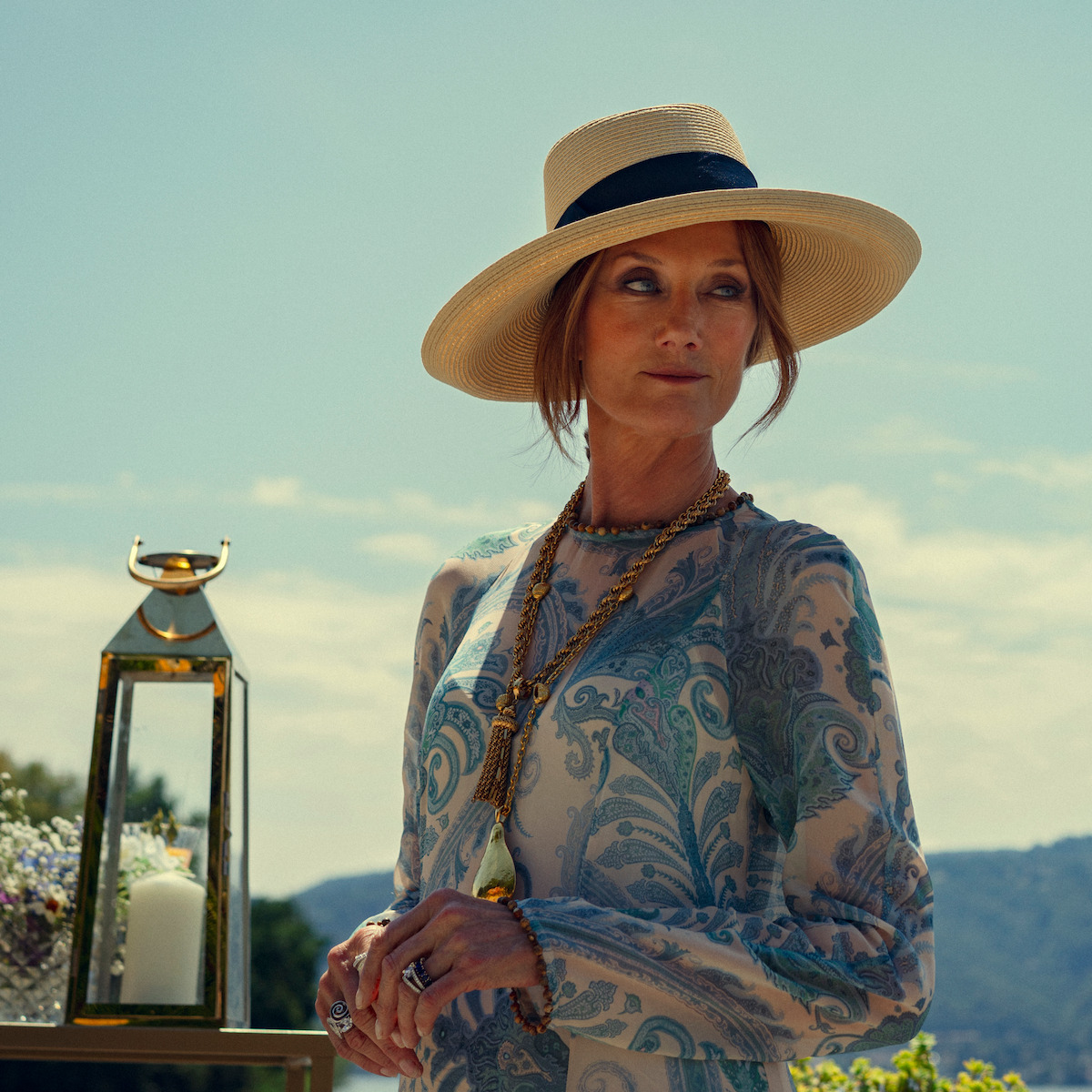 Woman in a paisley dress and straw hat standing outdoors on a sunny day, wearing layered necklaces, with a decorative lantern and candle beside her, mountains and blue sky in the background.