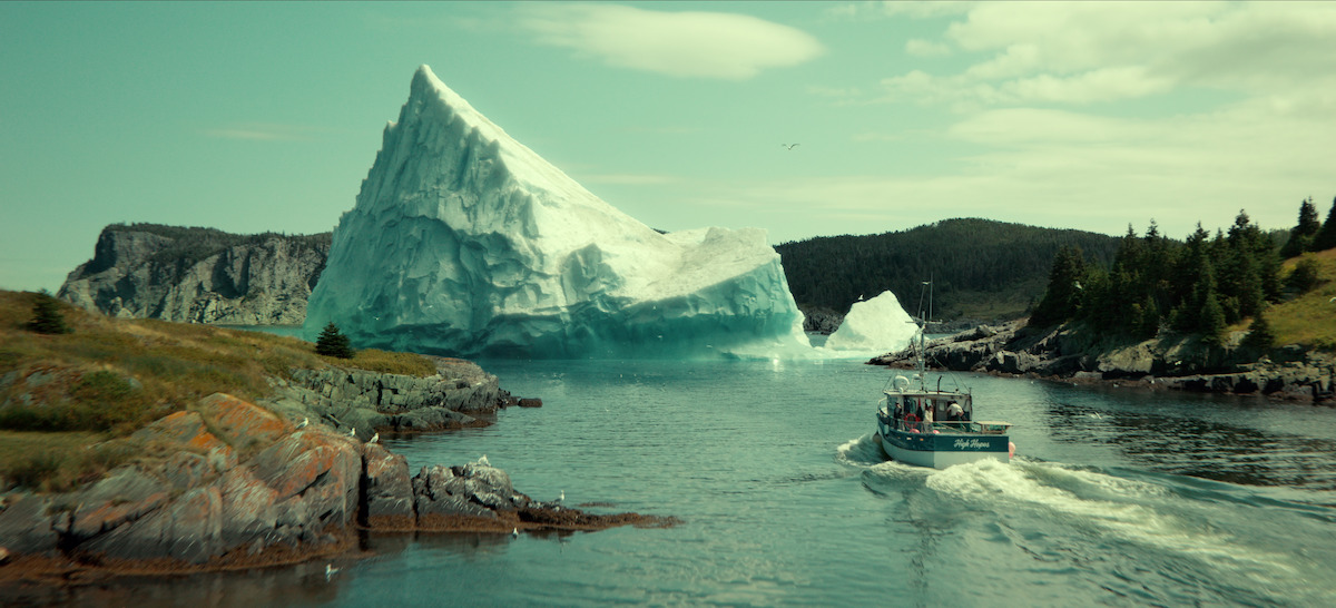 Fishing boat sailing through a narrow waterway flanked by rocky shores and evergreen trees, with a massive iceberg floating ahead, under a partly cloudy sky.