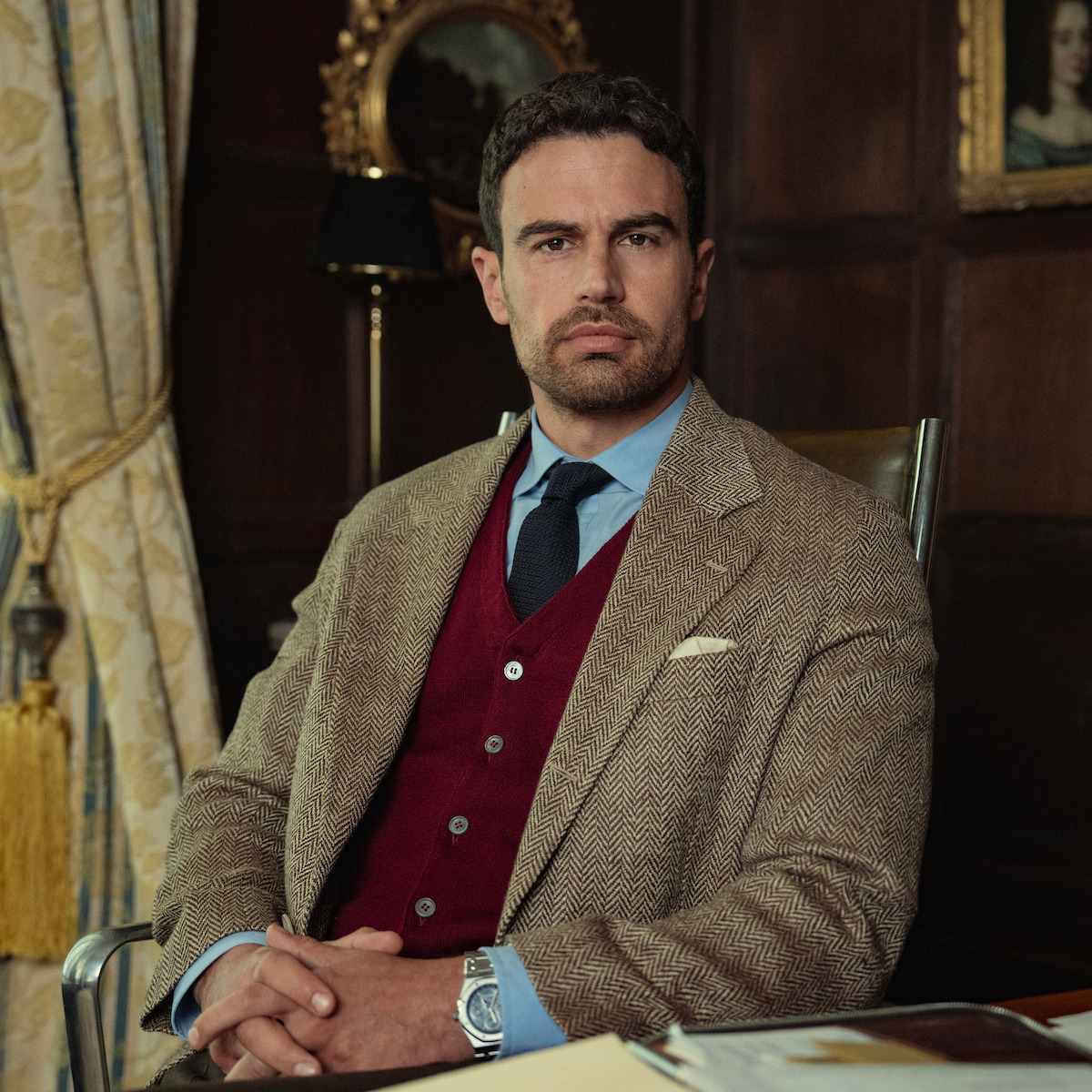 Man in a suit and red vest sitting at a desk in a formal office with wood-paneled walls, ornate curtains, framed art, and documents on the desk, looking serious and composed.