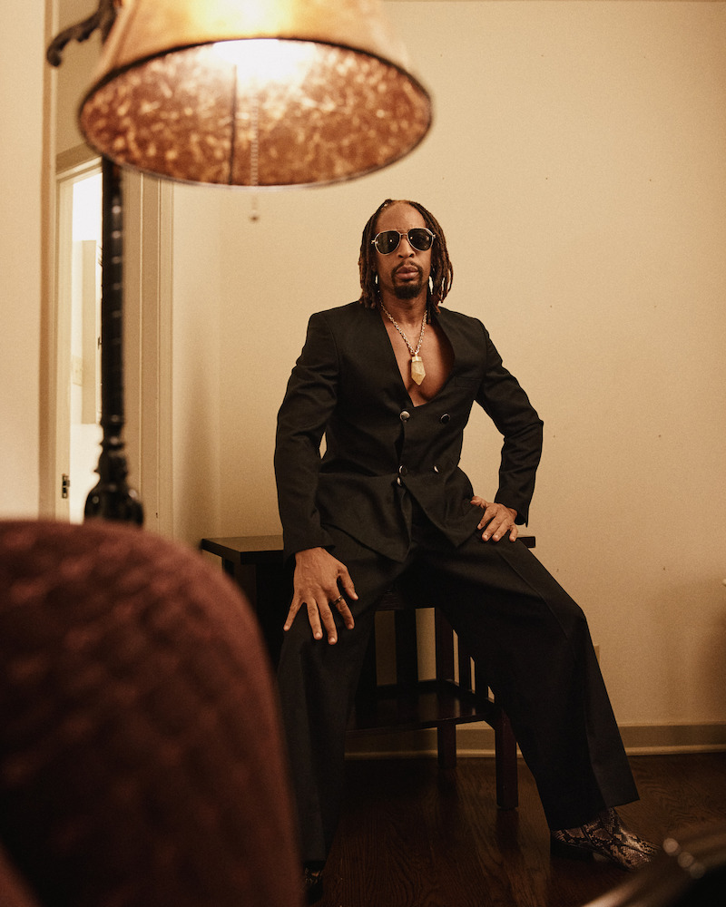 A man in a black suit and sunglasses poses confidently in a dimly lit room with wood floors, a lamp in the foreground, and neutral-colored walls and furniture.