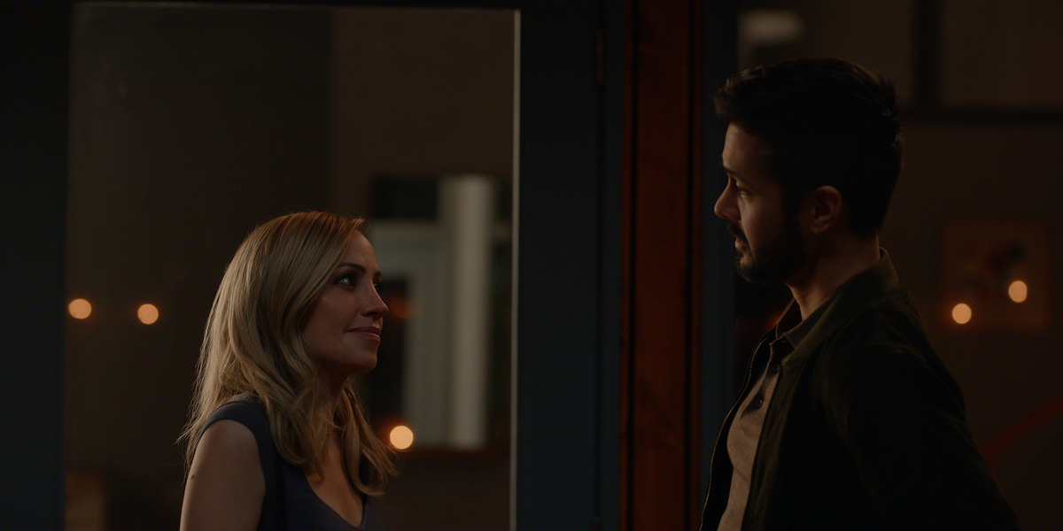 A woman and a man stand facing each other in a dimly lit indoor setting, possibly having a conversation at night, with warm lights softly illuminating the background.