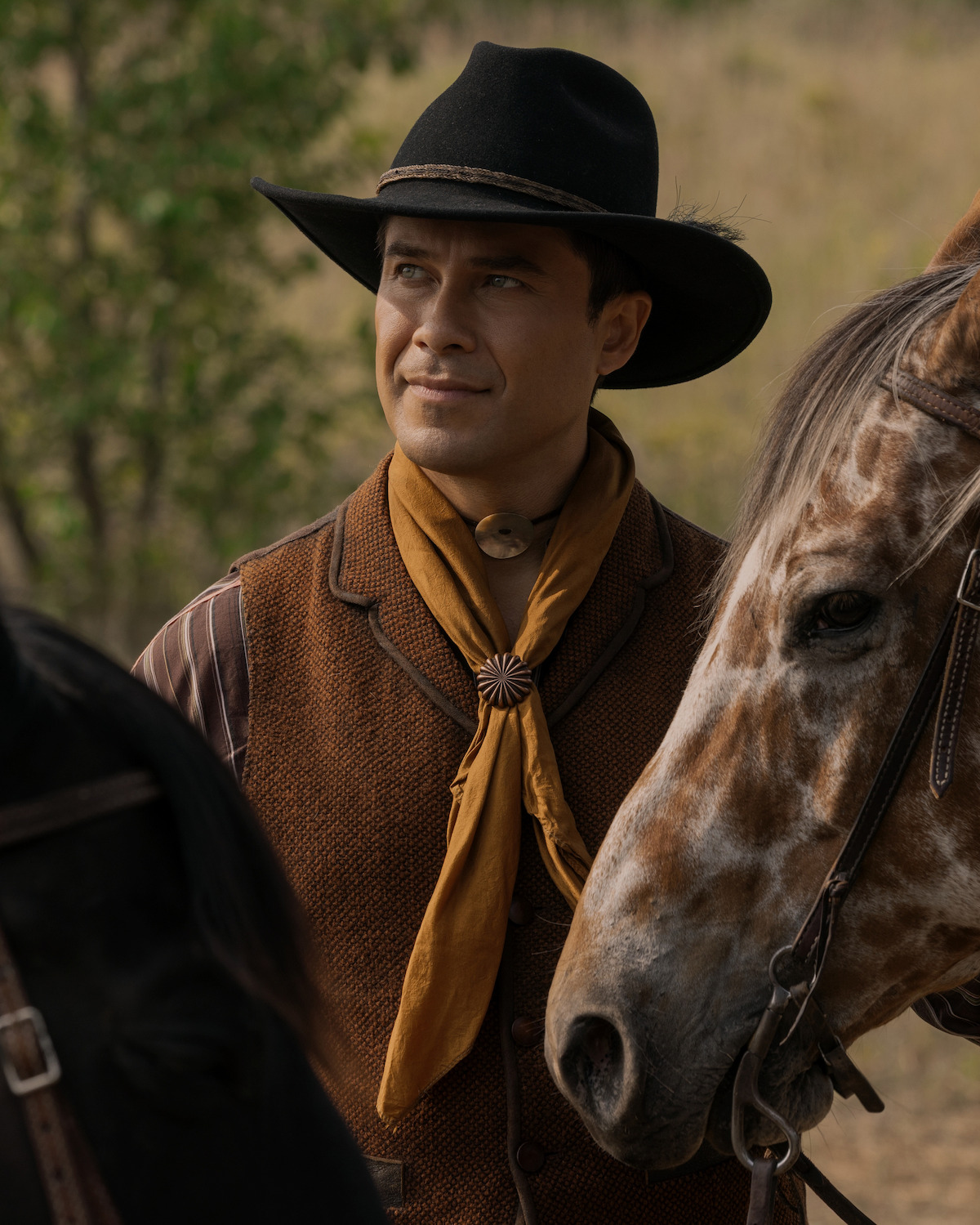 Man in cowboy attire with a black hat and yellow scarf stands next to a brown and white horse outdoors in a natural, grassy setting with blurred trees in the background.