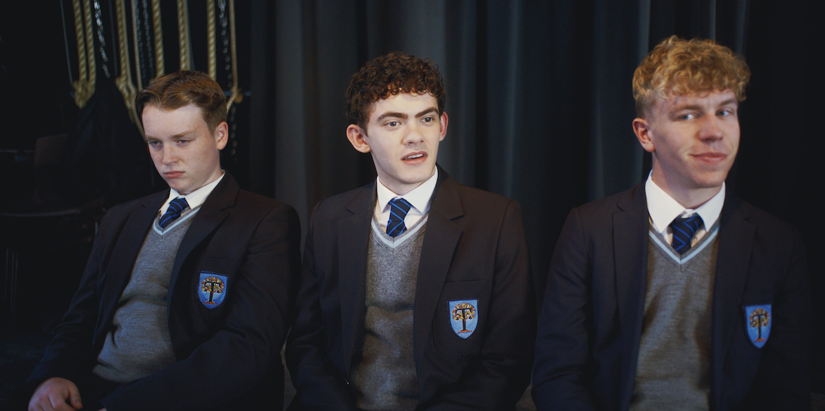 Three male students in school uniforms sit together on a bench indoors, with dark curtains and ropes in the background, appearing to be in a formal or staged setting, each displaying different facial expressions.
