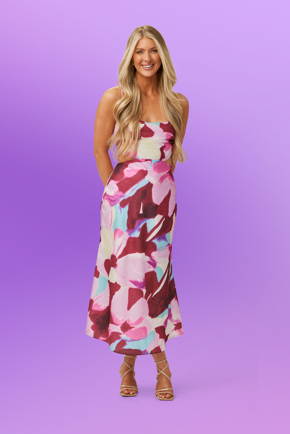 Ashley wearing a colorful floral dress and sandals, standing in front of a smooth purple gradient background in a studio setting.