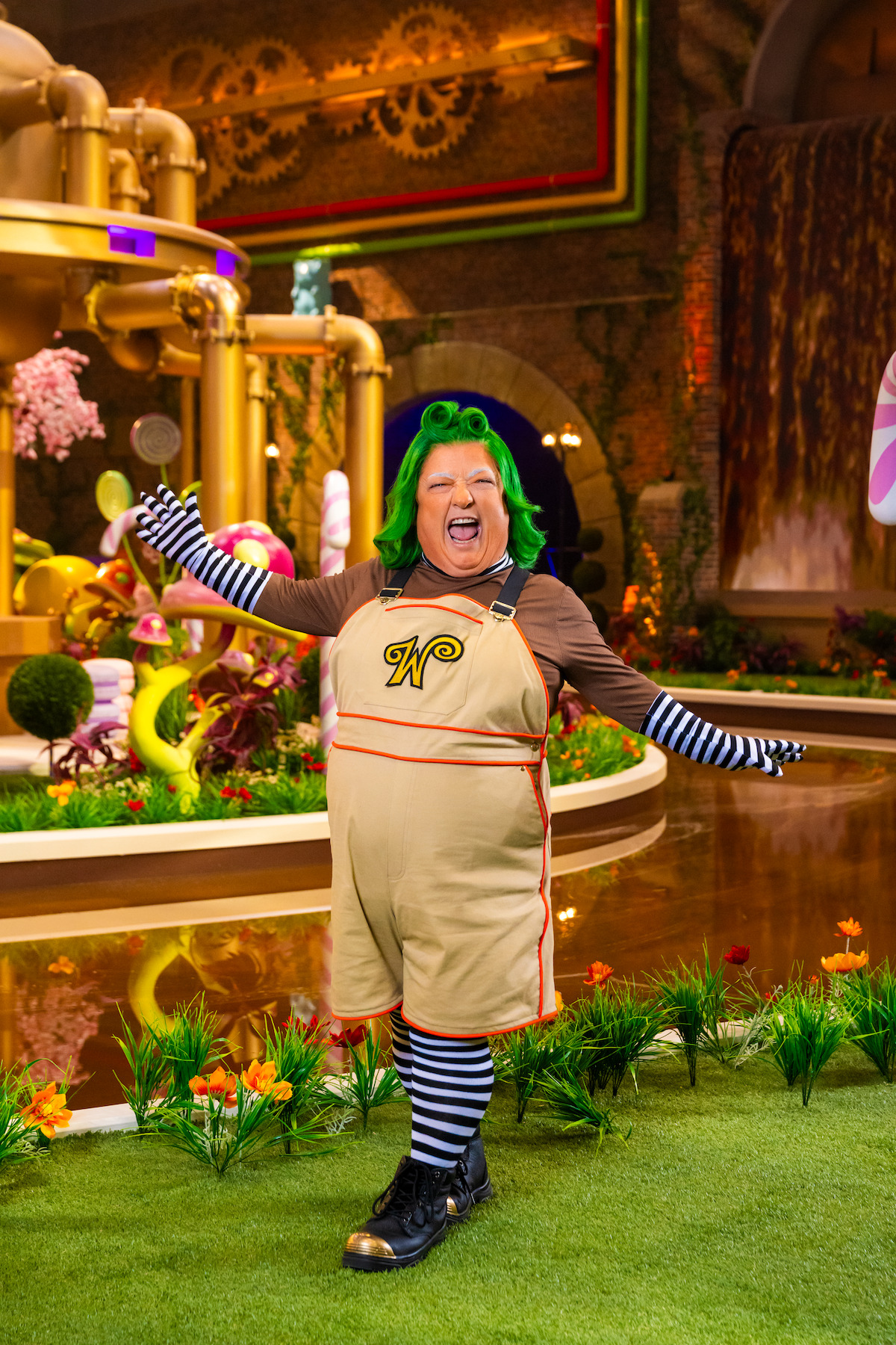 Person dressed as an Oompa Loompa with green hair, striped sleeves and socks, standing joyfully in a whimsical, colorful candy factory set with pipes, candy decorations, and lush greenery.