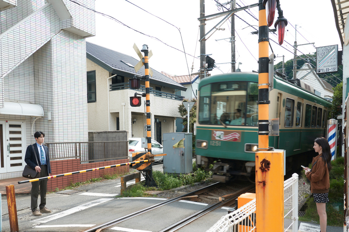 A green train crossing a small urban railway in a residential neighborhood, with a man and woman waiting at the crossing gate, surrounded by houses and utility poles.