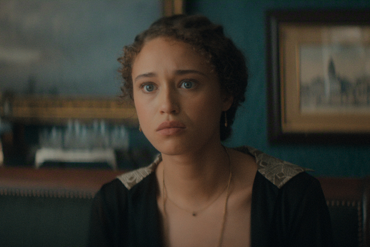 A young woman with curly hair sits indoors, looking thoughtful or concerned. She is in a dimly lit room with framed paintings and antique decor in the background.