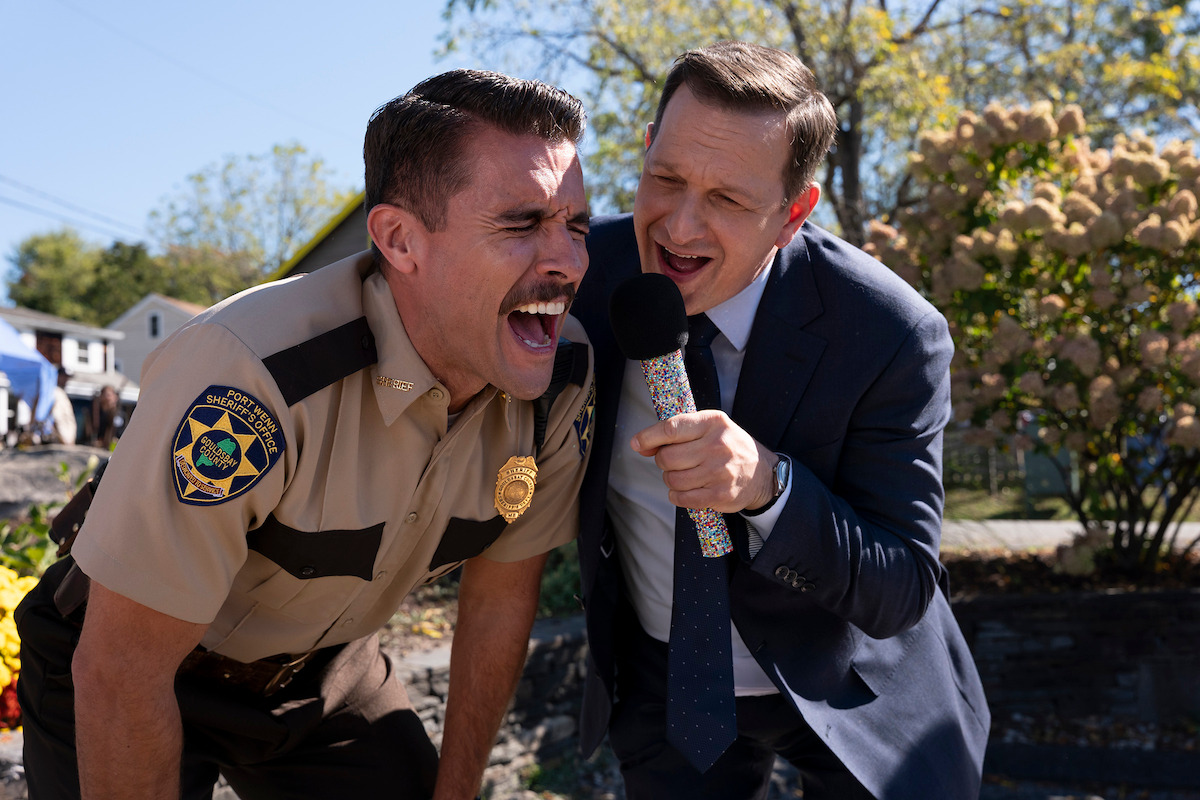 A police officer in uniform leans forward and yells while a man in a suit enthusiastically holds a microphone up to him outdoors, with trees and houses in the background on a sunny day.