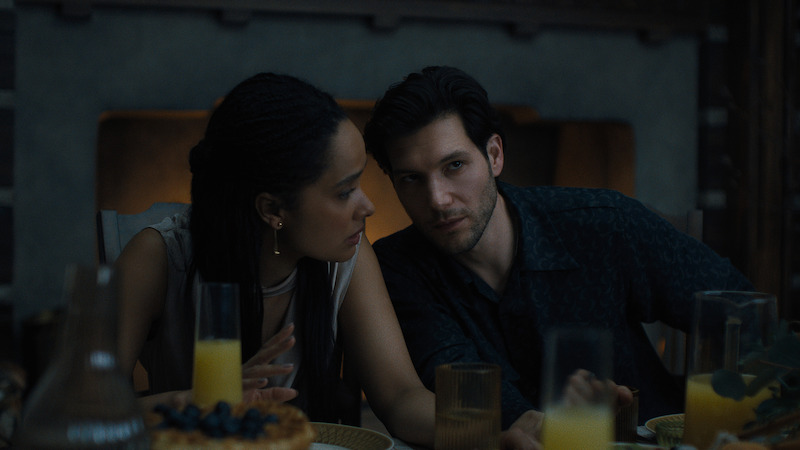 A woman and man sit closely at a dimly lit dinner table with breakfast foods, juice, and candles, suggesting an intimate conversation in a cozy indoor setting.