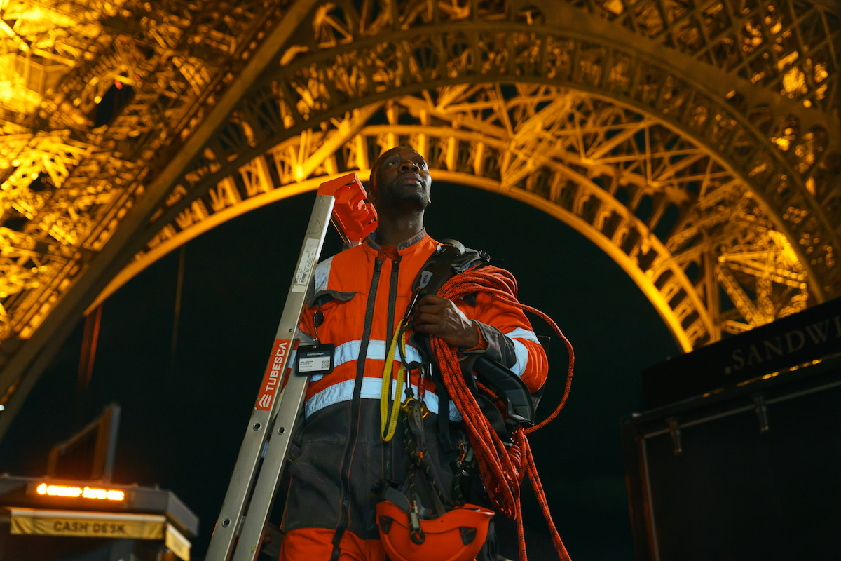 A worker in orange safety gear stands under the illuminated Eiffel Tower at night, holding a ladder and rope, with dark sky and structural metal details visible above.