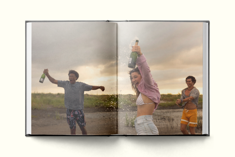 Three young adults outdoors in a grassy, open area near water, celebrating and spraying champagne, smiling and wearing summer clothes with cloudy skies in the background, captured in a photo book.