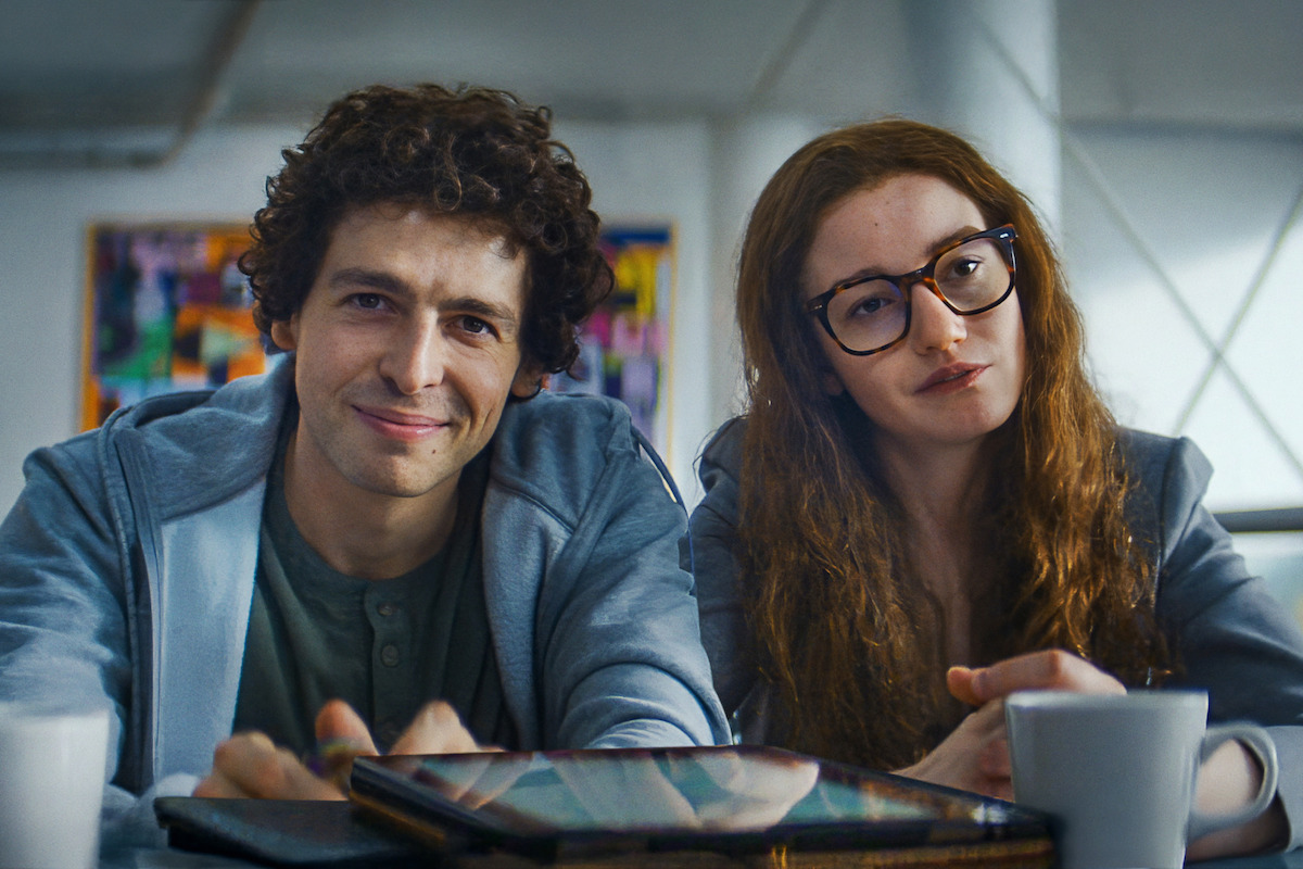 Two young adults sitting side by side at a table in a bright, modern indoor setting with art on the wall, coffee cups, and tablets in front of them.
