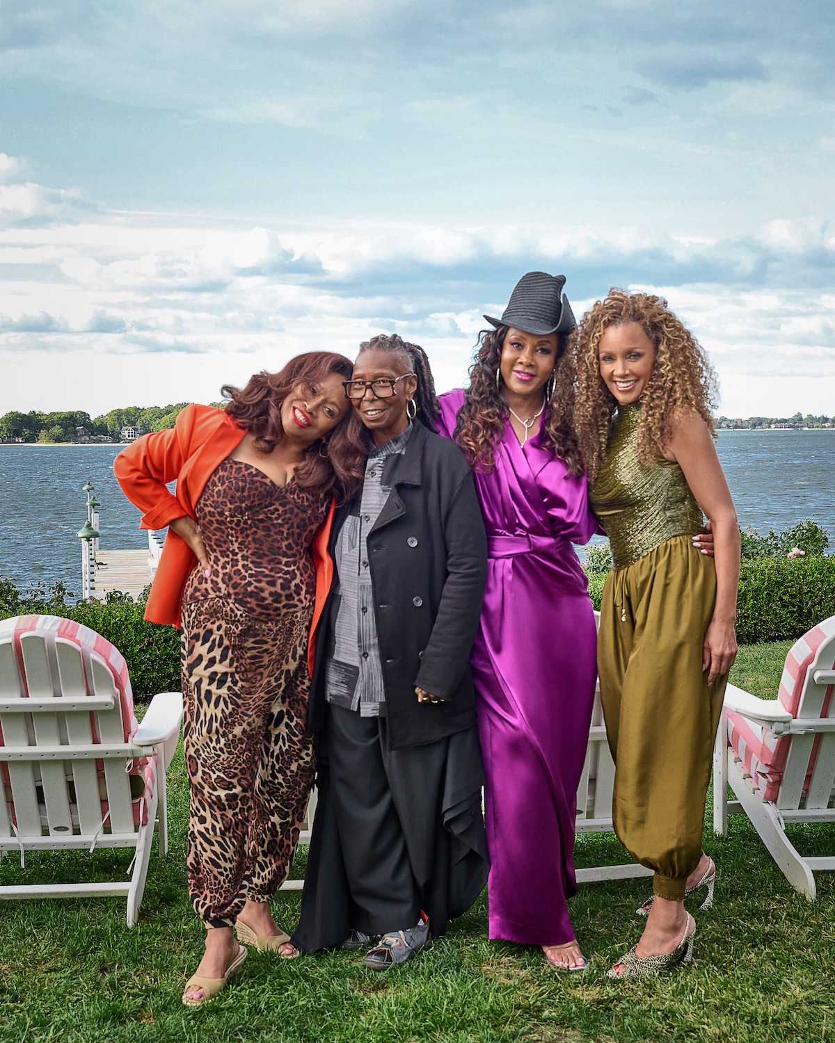 Four women posing together on a grassy lawn in front of a lake, with white chairs and green shrubs in the background. They are dressed in vibrant, stylish outfits, smiling and enjoying an outdoor gathering on a partly cloudy day.