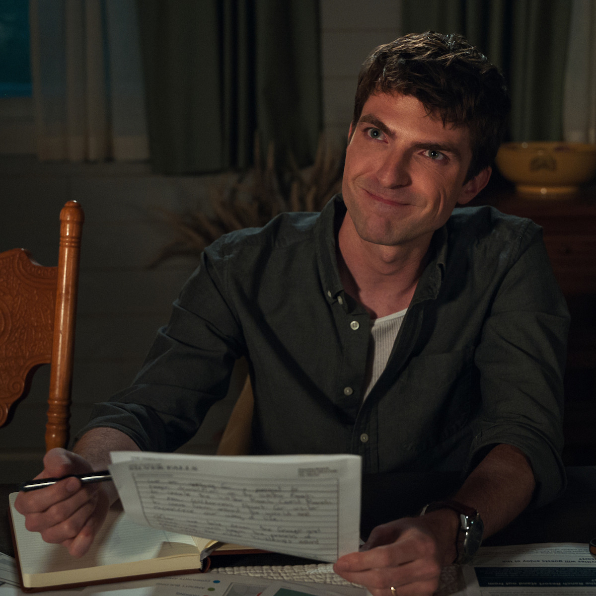 Smiling man sitting at a wooden table in a cozy, dimly lit room, holding a document and pen. Open notebook and paperwork are on the table, with a chair and curtains in the background.